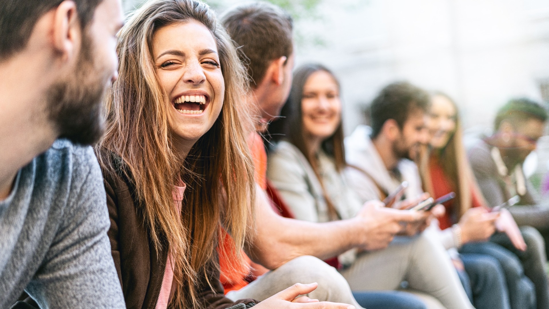 Eine Gruppe trendiger junger Leute, die auf einer Bank im Freien sitzen und sich unterhalten. Studenten, die zusammen Spaß haben. Fokus auf ein blondes Mädchen, das mit offenem Mund lächelt